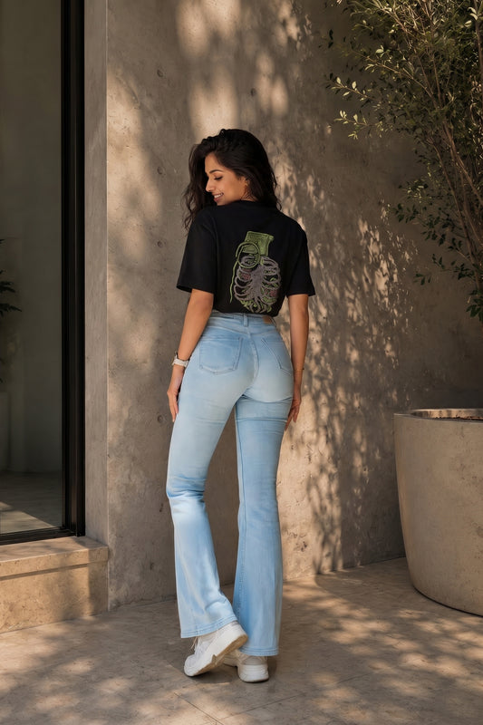 Person wearing a black t-shirt with an embroidery design and light blue jeans standing against a beige wall.