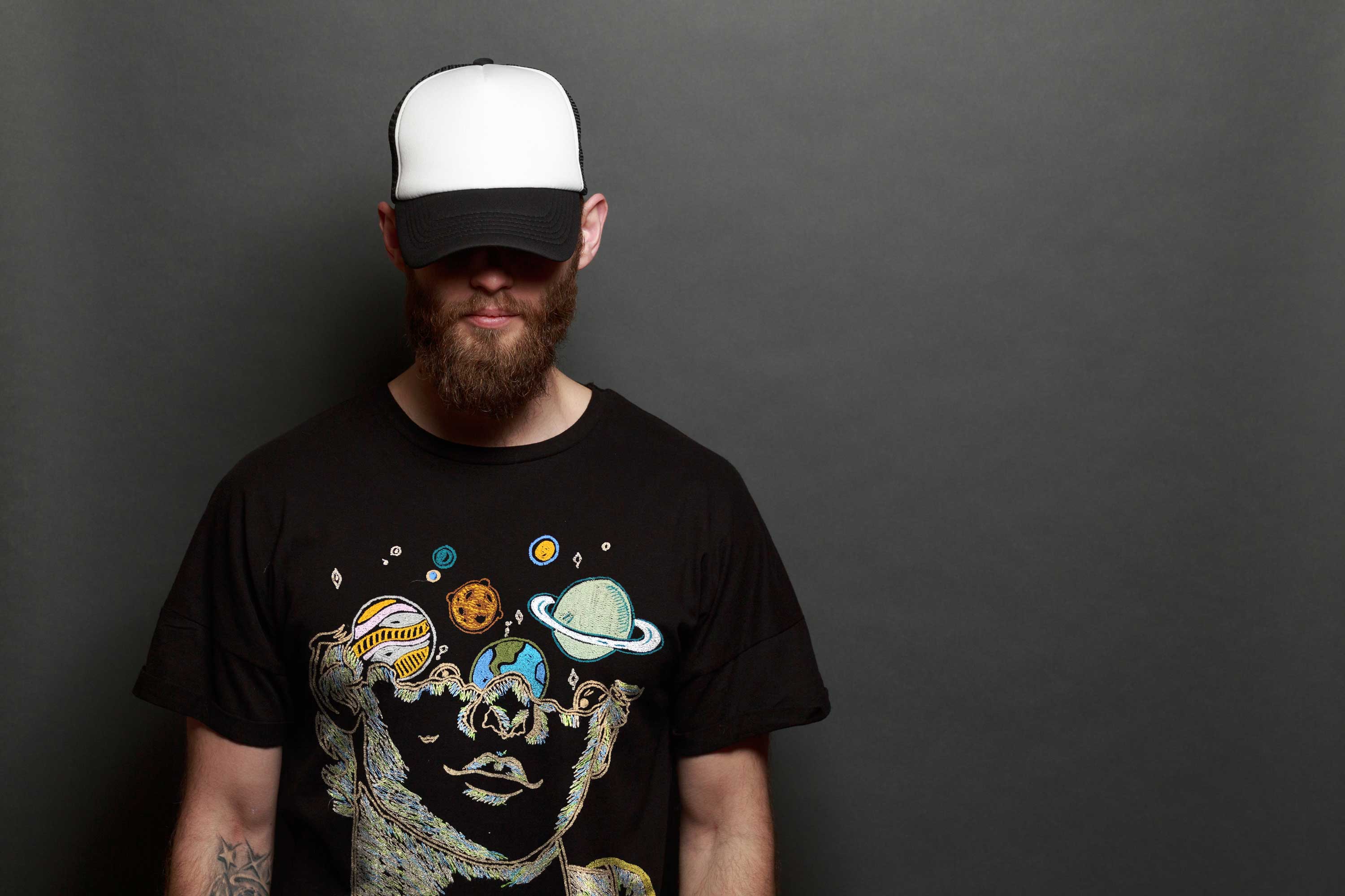 Man wearing a black t-shirt with space-themed design and a white cap on a dark background