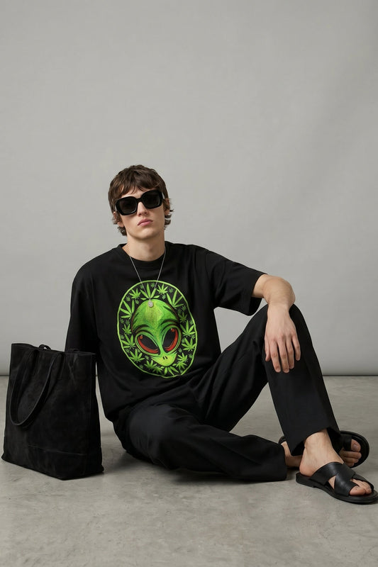 Person wearing a black t-shirt with an alien design, sitting on the floor against a gray background.