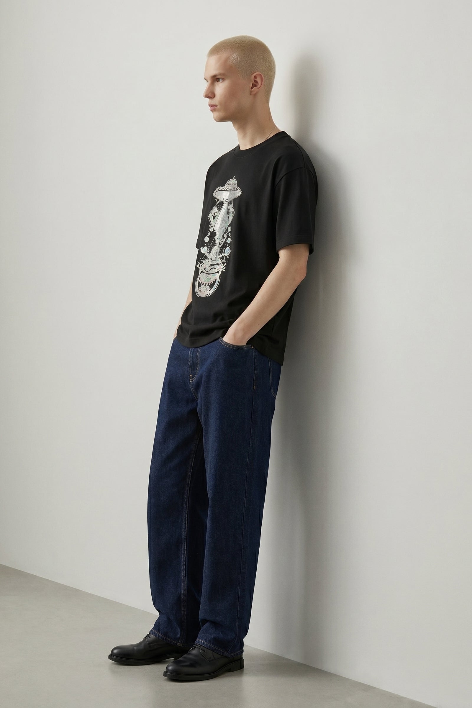 Person wearing a black t-shirt with a graphic design and blue pants against a white wall.