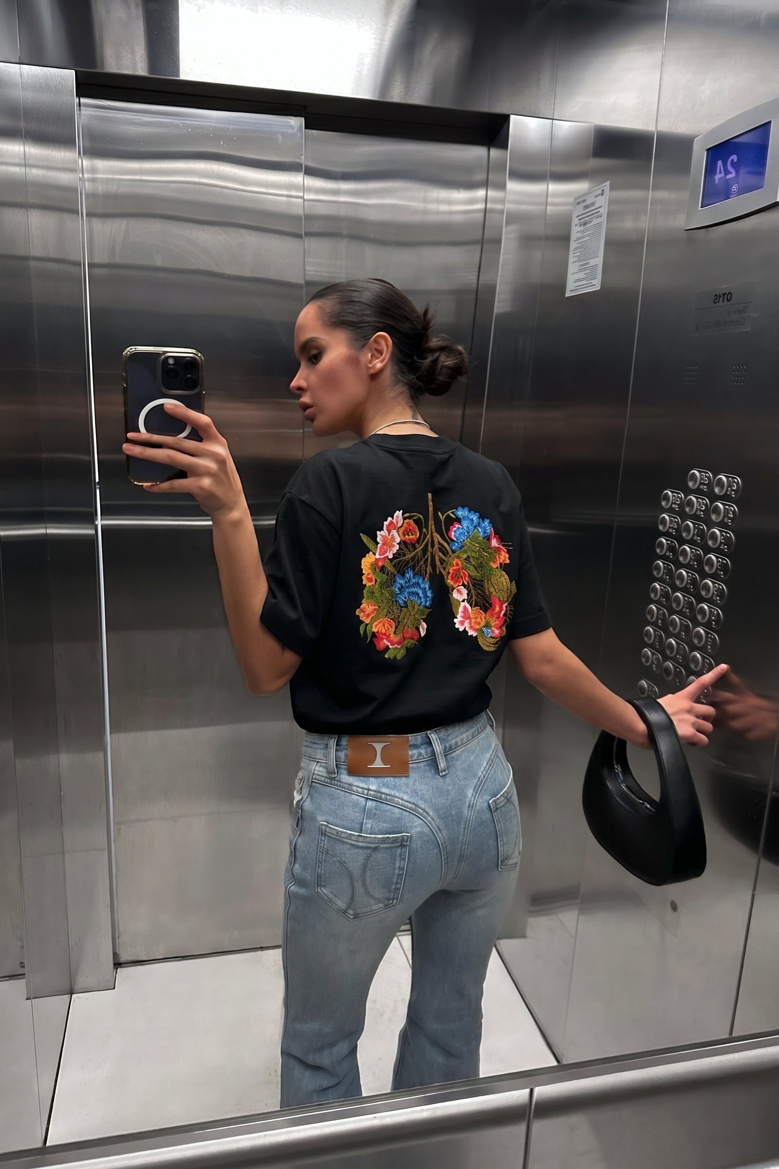 Person taking a mirror selfie in an elevator wearing a black t-shirt with colorful floral design.