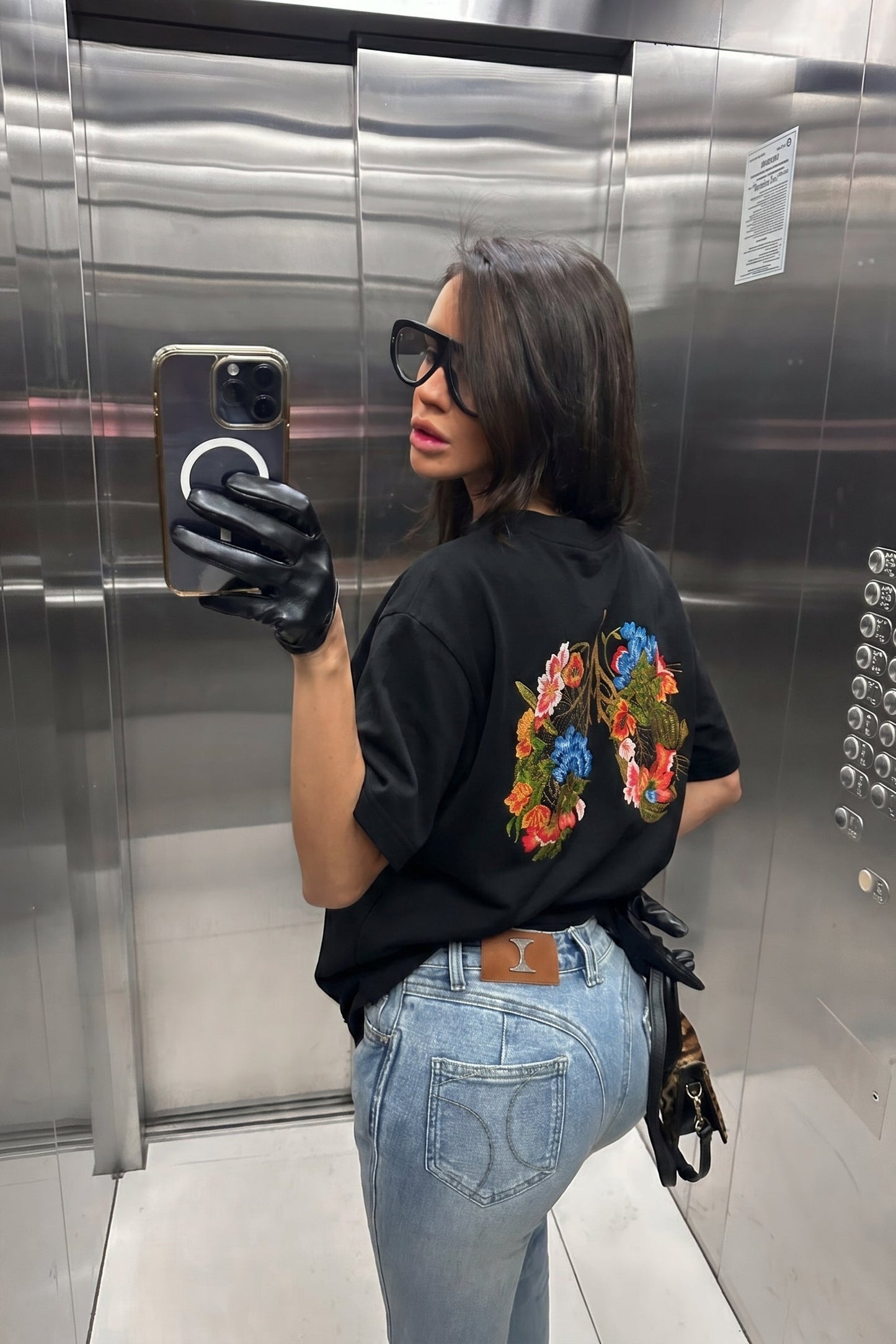 Person taking a mirror selfie in an elevator wearing a black t-shirt with colorful designs and blue jeans.