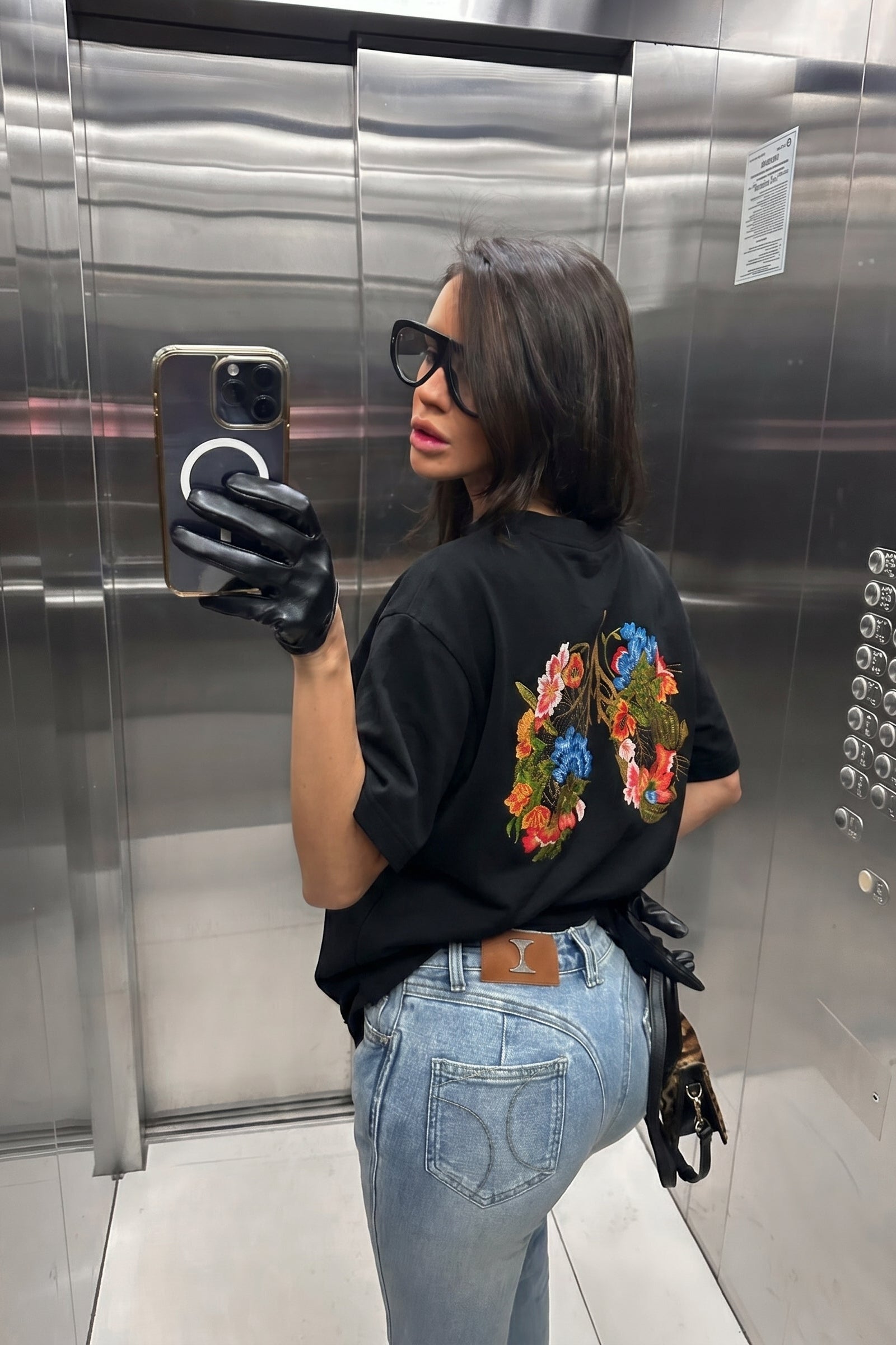 Person taking a mirror selfie in an elevator wearing a black t-shirt with colorful designs and blue jeans.