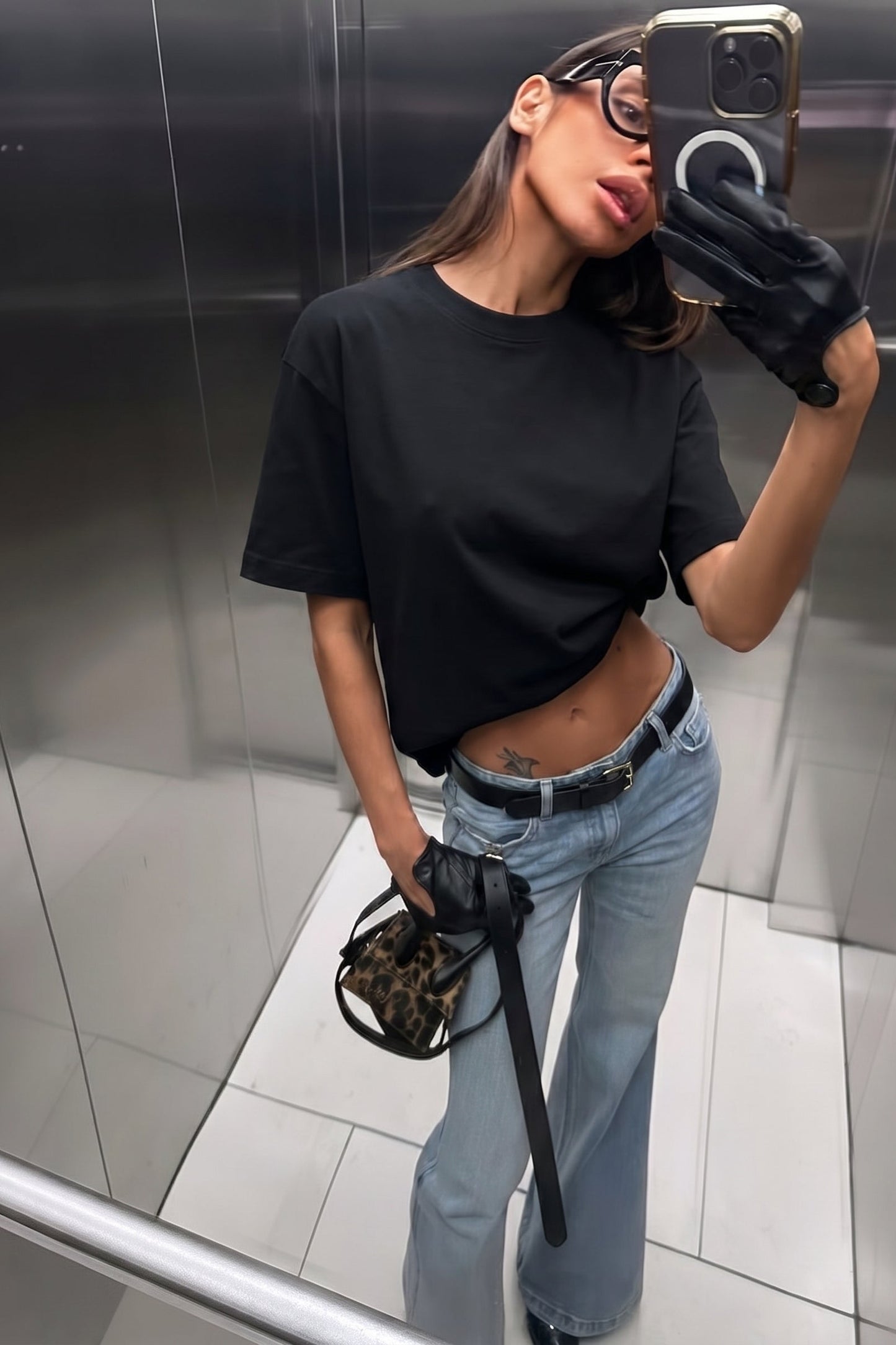 Person taking a mirror selfie wearing a black t-shirt, blue jeans, and black gloves in an elevator.