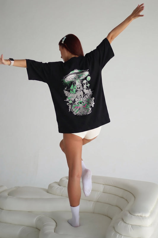 Person wearing a black oversized t-shirt with a colorful graphic design, standing on a white couch against a plain background.