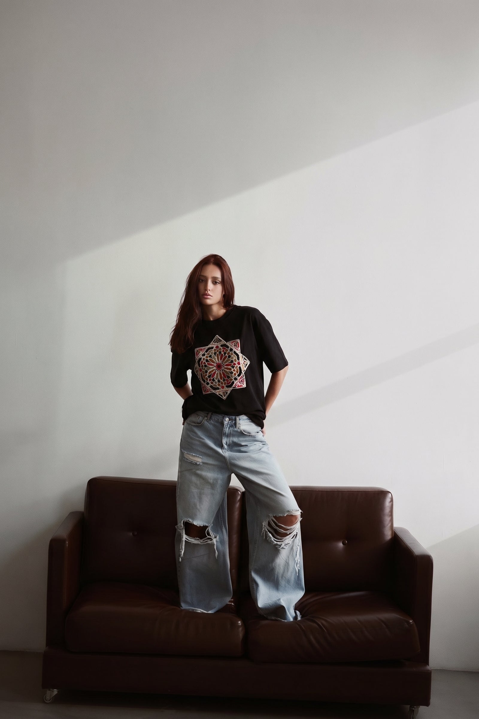 Person wearing a black t-shirt with a colorful design and light blue jeans standing on a brown leather couch against a white wall.