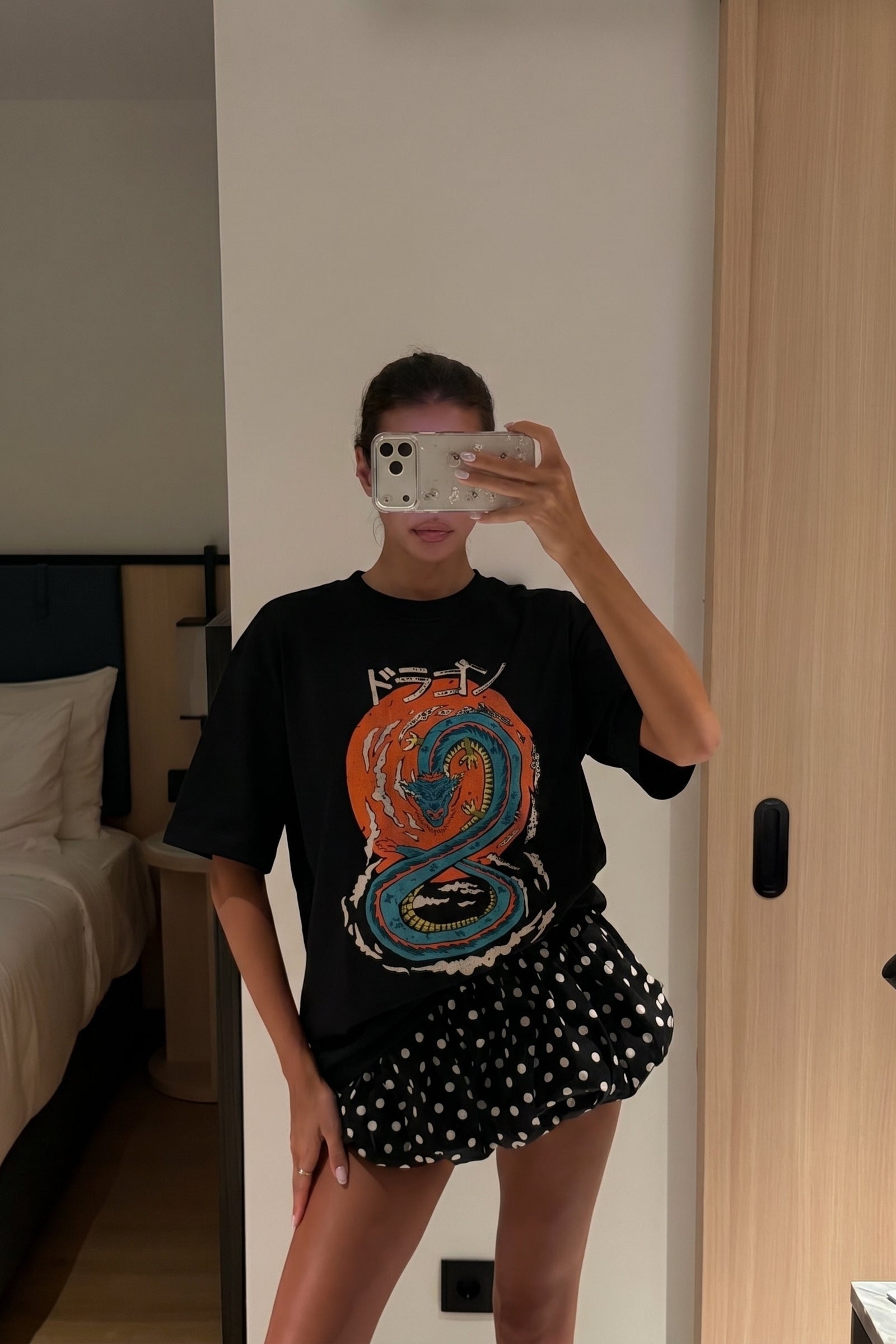Person taking a mirror selfie wearing a black t-shirt with a colorful design and a polka dot skirt.
